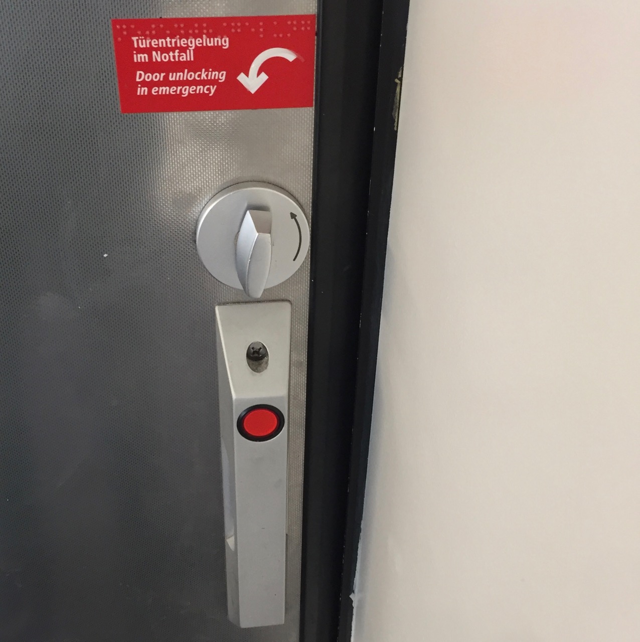 The inside of the toilet door, a similar door handle as before is visible. But this time the red circle is at the top, and there is no green area for opening or closing. There is only a weird mechanical dial above the handle. And above that dial is a white note on red ground that says "Door unlocking emergency", next to an arrow indicating that dial must be turned counter-clockwise.