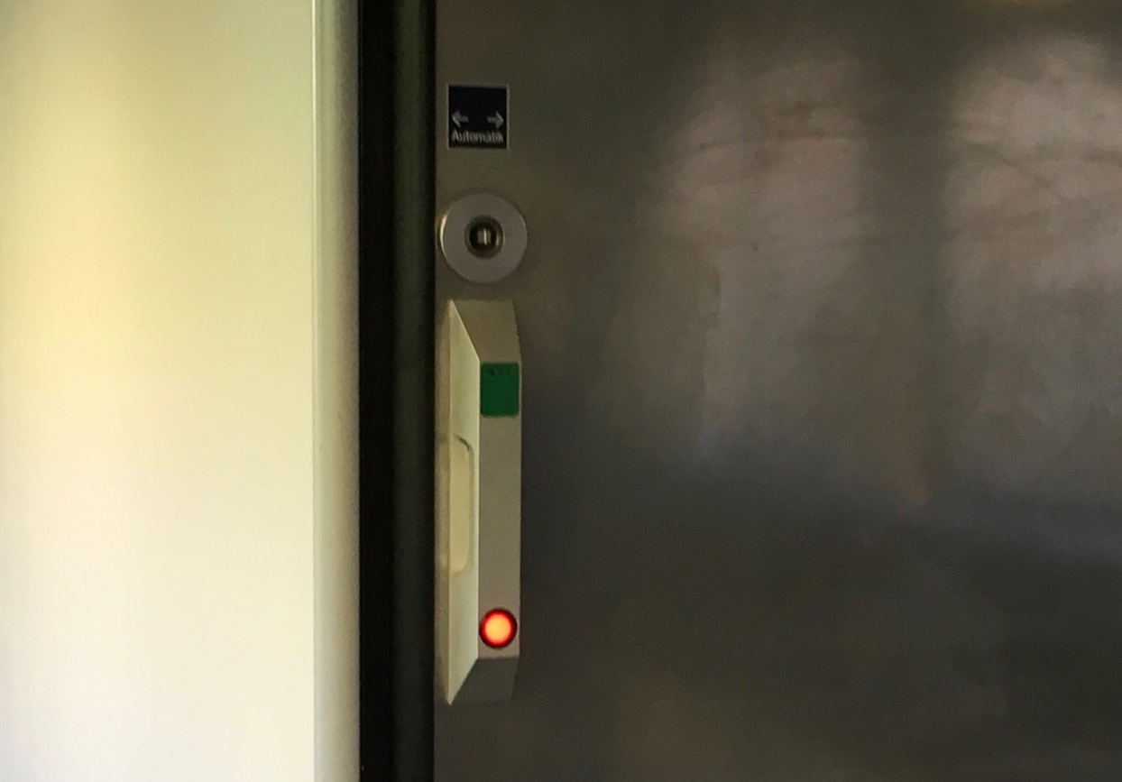 The "controls" for a the disability-friendly toilets in german trains. Visible is a small vertical bar, which looks a bit like a handle, attached to a metal sliding door. At the top end of the bar there is a green rectangle, at the bottom there is a shining red circle with a black border.